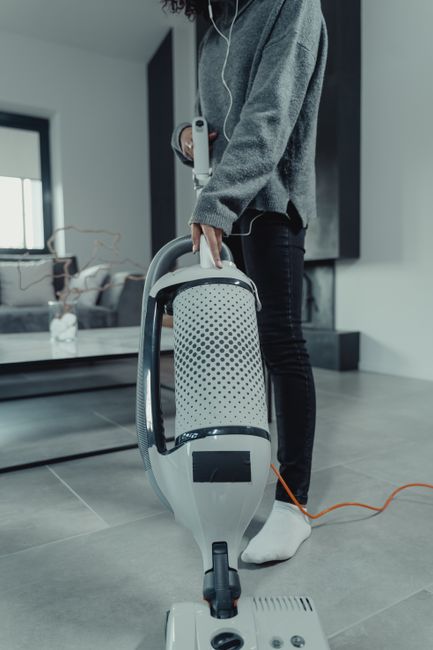 A woman with curly hair, wearing a green jacket, blue jeans, and white shoes, is using a yellow vacuum cleaner with a hose attachment to clean a patterned area rug in a living room. The room features 